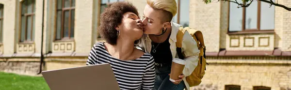 Two young women share a tender kiss while holding laptop in front of a vibrant building on campus. — Stock Photo