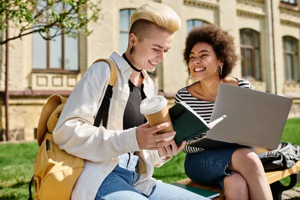 Due giovani donne in abbigliamento casual siedono su una panchina, ognuna delle quali lavora su un computer portatile, focalizzata e impegnata nei loro compiti. — Foto stock