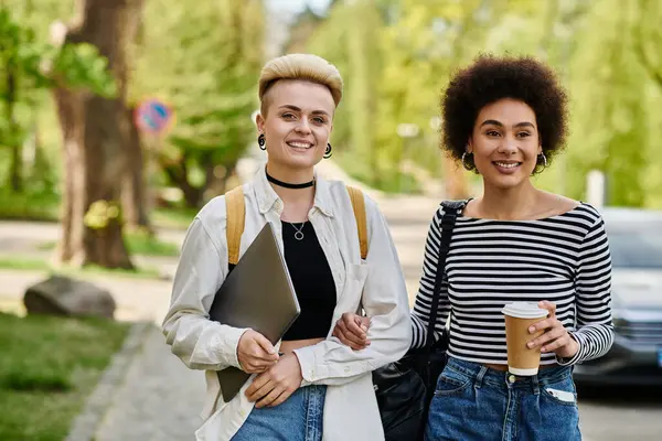 Zwei junge Frauen, eine schwarze und eine weiße, gehen mit Kaffeetassen die Straße hinunter und unterhalten sich fröhlich. — Stockfoto