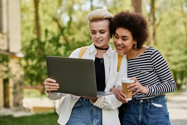 Two young women, casually dressed, sit side by side in a park, focused on a laptop screen, engaged in a conversation. — Stock Photo
