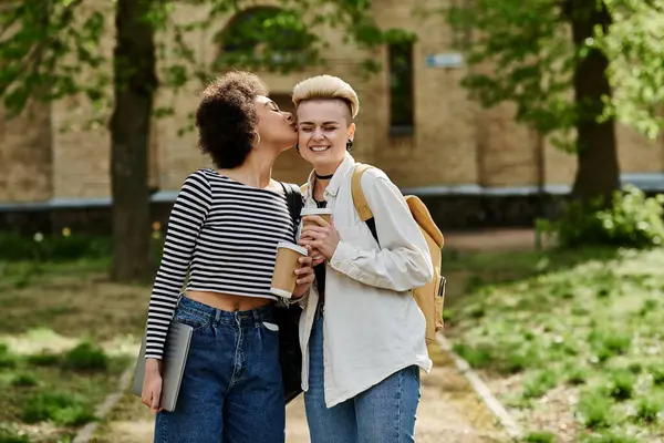 Two young women in stylish attire sharing a tender kiss amidst lush greenery in the park. — Stock Photo