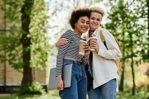 Two young women, holding coffee and a laptop, sit in a park surrounded by nature, focused on their work. — Stock Photo