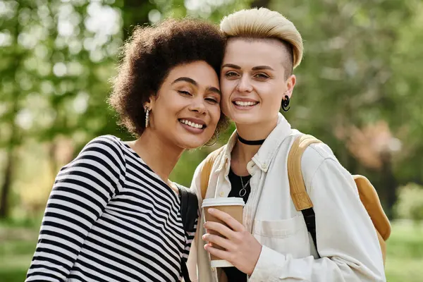 Deux jeunes femmes, élégamment habillées, tiennent joyeusement des tasses à café dans un parc animé près d'un campus universitaire. — Photo de stock