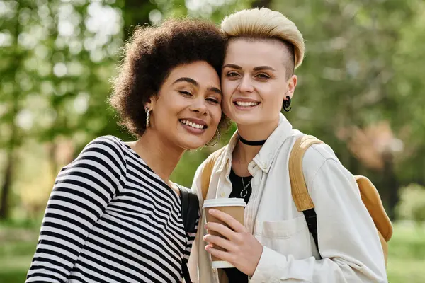 Zwei junge Frauen, stilvoll gekleidet, halten fröhlich Kaffeetassen in einem lebhaften Park in der Nähe eines Universitätscampus. — Stockfoto