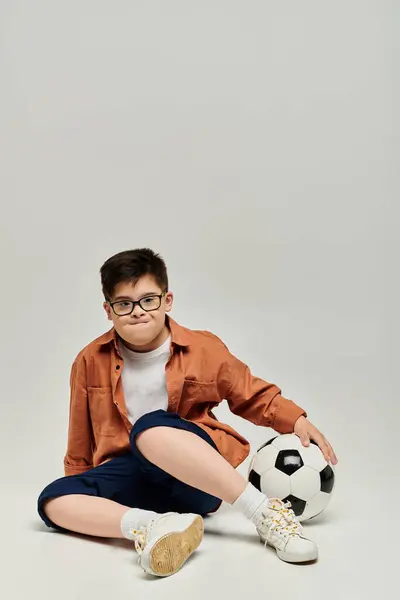 Menino com síndrome de Down com óculos senta-se no chão com bola de futebol. — Fotografia de Stock