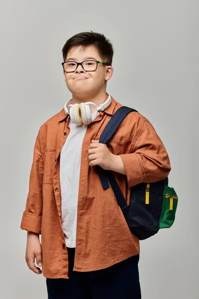 Un niño pequeño con síndrome de Down con gafas y una mochila mirando a su alrededor con curiosidad. - foto de stock