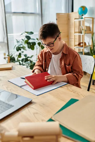 Un niño con síndrome de Down se sienta en un escritorio con una computadora portátil y un libro. - foto de stock