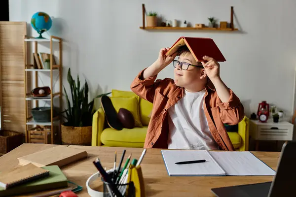 A boy with Down syndrome sitting at a desk, balancing a book on his head. — Stock Photo