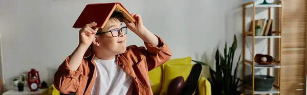 Little boy with Down syndrome holds a book over his head while sitting on a couch. — Stock Photo