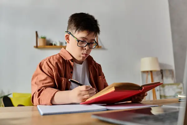 A boy with Down syndrome reads a book and works on a laptop at a desk. — Stock Photo