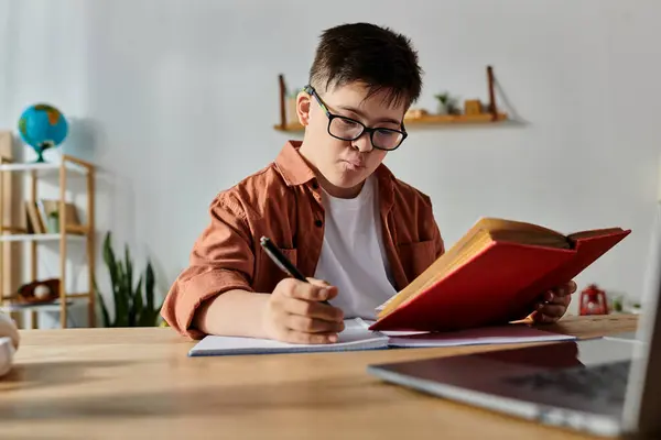 A boy with Down syndrome sits at a desk with a laptop and a book. — Stock Photo