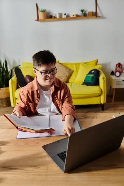 Un garçon trisomique assis à une table avec un ordinateur portable et un livre. — Photo de stock