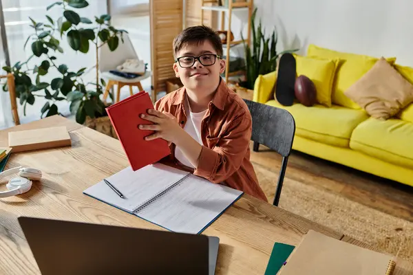 A boy with Down syndrome seated at a desk, focused on his notebook and laptop. — Stock Photo