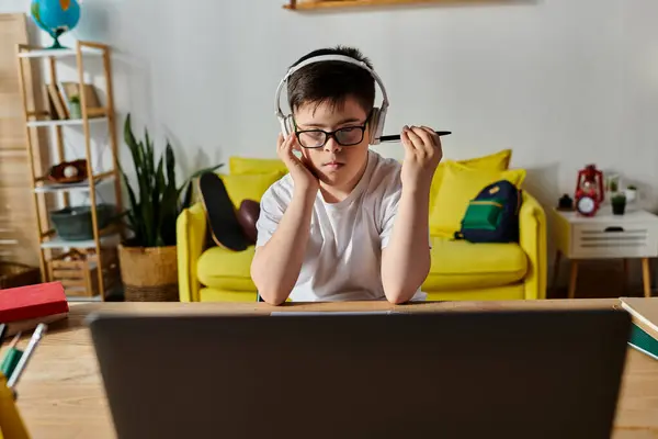 A boy with Down syndrome sits at a table with headphones, focusing on his laptop. — Stock Photo