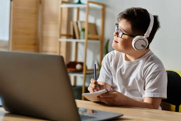 A adorable boy with Down syndrome wearing headphones sits at a desk using a laptop. — Stock Photo