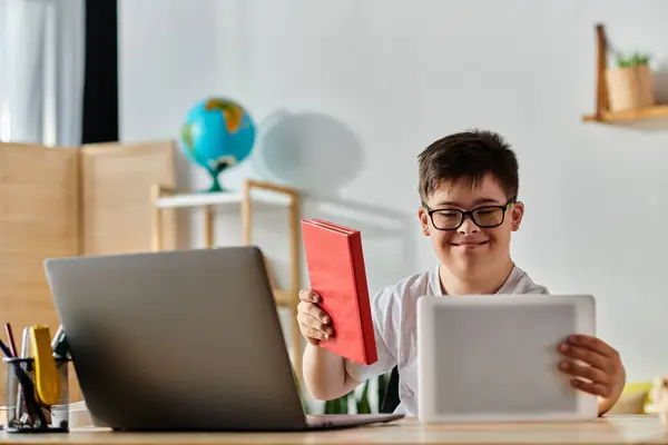 Um menino com síndrome de Down sentado em uma mesa com um laptop e livro. — Fotografia de Stock