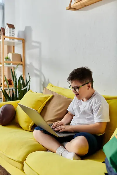 A boy with Down syndrome uses a laptop on a bright yellow couch. — Stock Photo