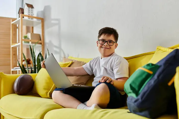 Un adorable garçon avec le syndrome de Down assis sur un canapé jaune, en utilisant un ordinateur portable. — Photo de stock