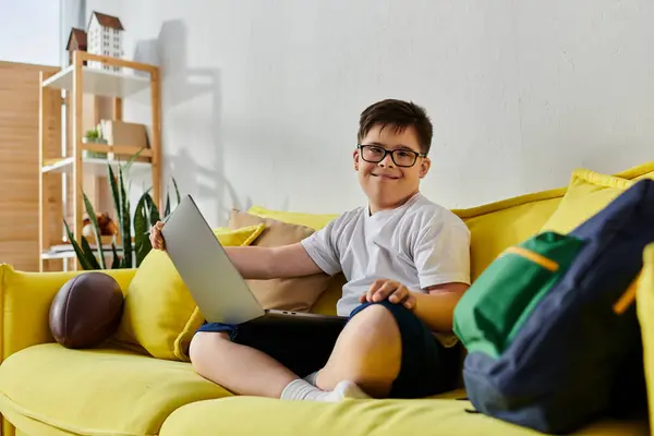 A adorable boy with Down syndrome sitting on a yellow couch, using a laptop. — Stock Photo