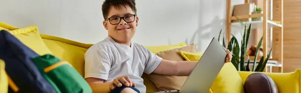 Adorable boy with Down syndrome sitting on yellow couch, using laptop. — Stock Photo