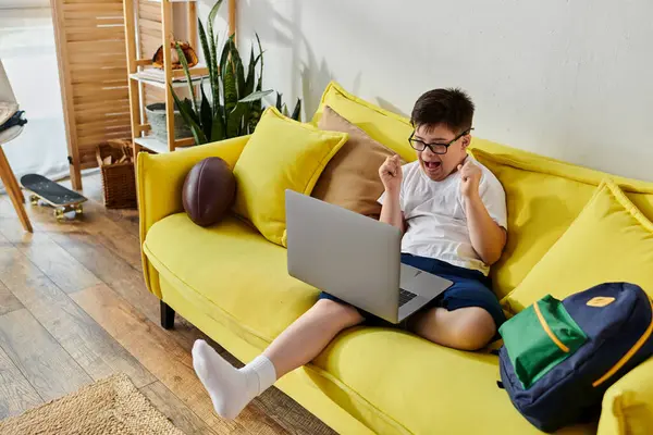 Un adorabile ragazzo con la sindrome di Down seduto su un divano giallo, utilizzando un computer portatile. — Foto stock