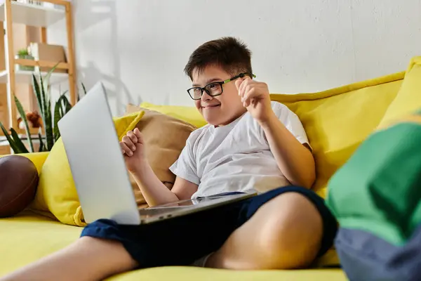 Adorable boy with Down syndrome sitting on yellow couch, using laptop. — Stock Photo