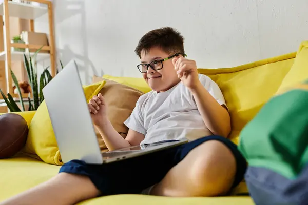 Adorabile ragazzo con sindrome di Down seduto sul divano giallo, utilizzando il computer portatile. — Foto stock