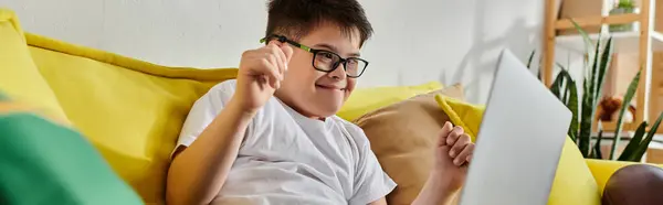 A boy with Down syndrome working on a laptop while seated on a yellow couch. — Stock Photo