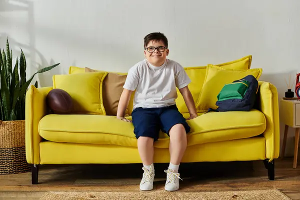 A cute boy with Down syndrome sitting on a bright yellow couch at home. — Stock Photo