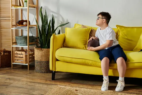 Ein entzückender Junge mit Down-Syndrom sitzt glücklich auf einer leuchtend gelben Couch in seinem Zimmer. — Stockfoto