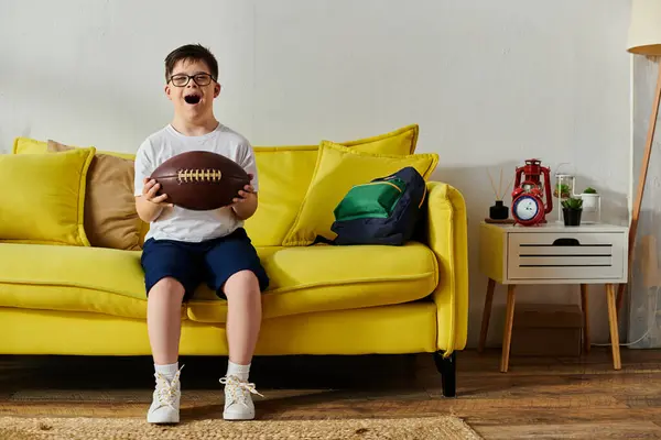A adorable boy with Down syndrome holding a football, seated on a bright yellow couch. — Stock Photo