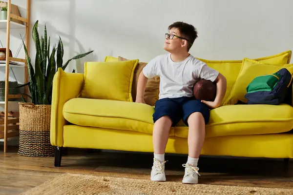 Un charmant garçon trisomique tenant un ballon de football assis sur un canapé jaune. — Photo de stock