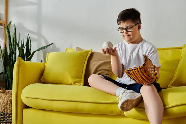 Um menino encantador com síndrome de Down repousa em um sofá amarelo ensolarado com uma luva de beisebol em suas mãos. — Fotografia de Stock