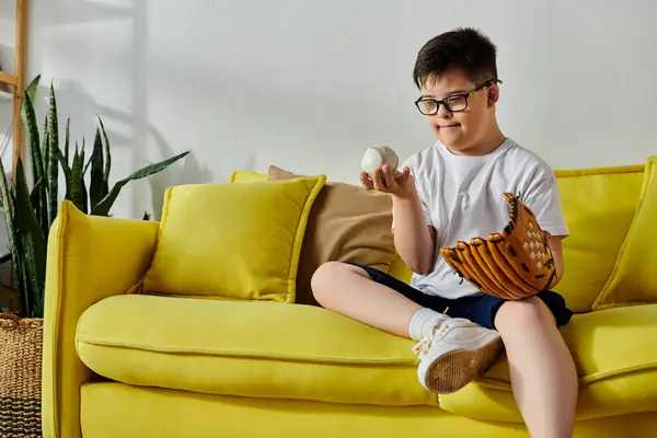 Um menino encantador com síndrome de Down repousa em um sofá amarelo ensolarado com uma luva de beisebol em suas mãos. — Fotografia de Stock