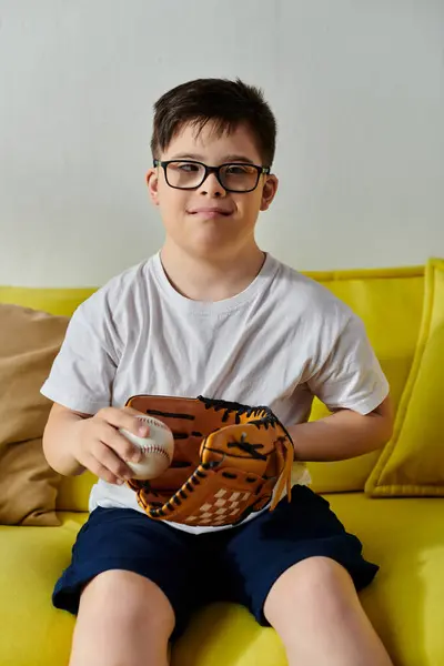 Adorable boy with Down syndrome with glasses sitting on a yellow couch holding a baseball. — Stock Photo