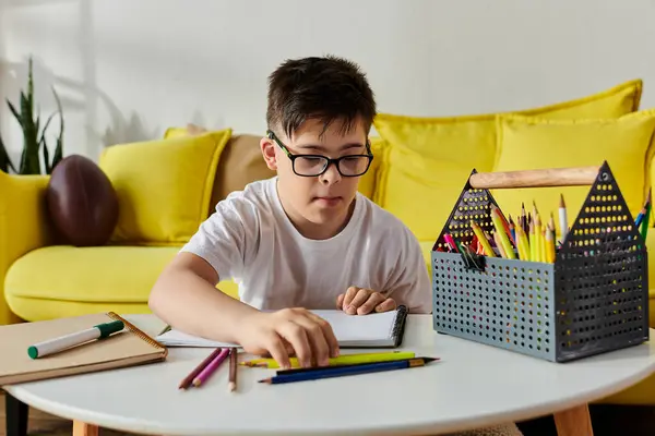 A little boy with Down syndrome sits at a table, engrossed in writing in his notebook with colored pencils. — Stock Photo