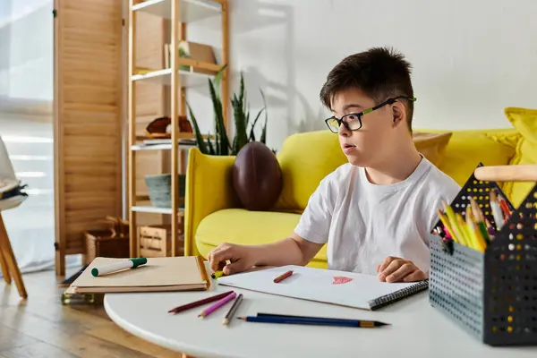 A boy with Down syndrome, wearing glasses, sits at a table focused on drawing. — Stock Photo