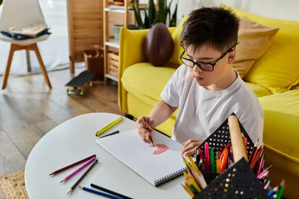 A adorable boy with Down syndrome sits at a table, engrossed in drawing with colored pencils. — Stock Photo