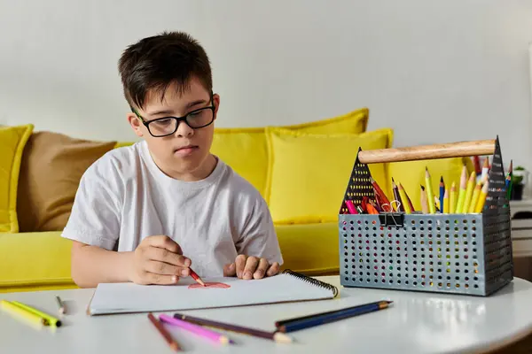 Мальчик с синдромом Дауна в очках рисует цветными карандашами за столом в своей комнате. — стоковое фото