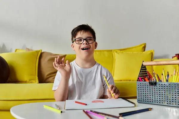 A adorable boy with Down syndrome in glasses happily drawing on a yellow couch. — Stock Photo