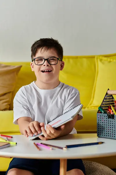 Adorable boy with Down syndrome with glasses at table, coloring in notebook with colored pencils. — Stock Photo