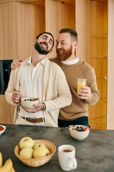 Feliz pareja gay se encuentra en la cocina moderna, compartiendo un tazón de fruta fresca. Tiempo de calidad y amor es evidente en sus sonrisas. - foto de stock