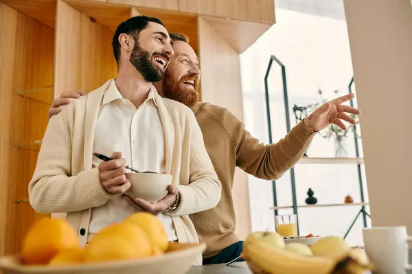 Two joyful men arranging a bowl of fresh fruit in a modern kitchen, sharing a loving moment together. — Stock Photo