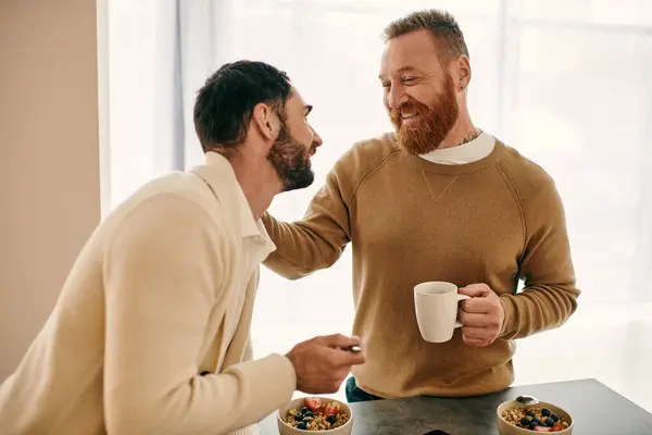 Dois homens, um feliz casal gay, estão sentados na mesa da cozinha, saboreando café e tempo de qualidade em seu apartamento moderno. — Fotografia de Stock