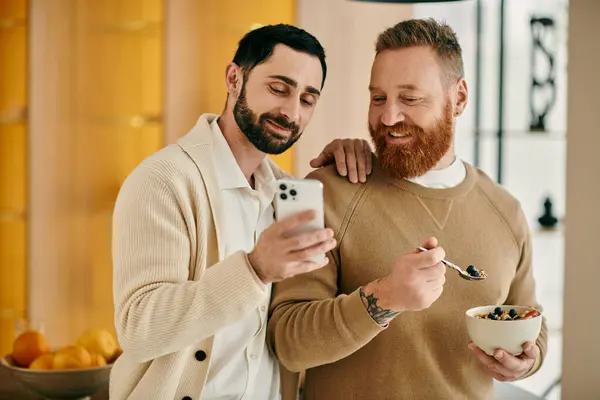 Two men, a happy gay couple, sit at a table eating breakfast while engrossed in phone — Stock Photo
