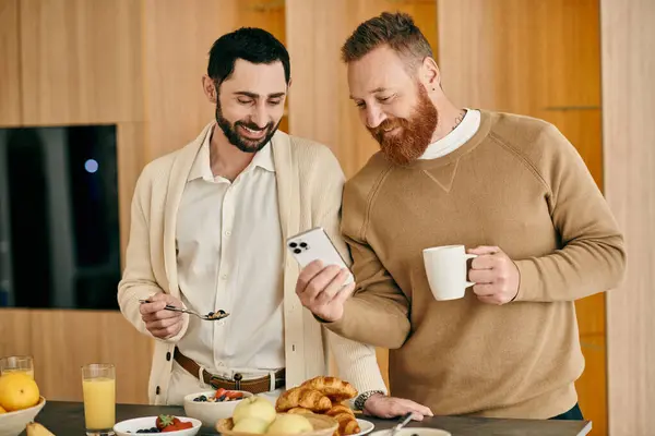 Dos hombres, uno con cabello oscuro y el otro con cabello claro, disfrutan del desayuno juntos mientras están absortos en el teléfono - foto de stock