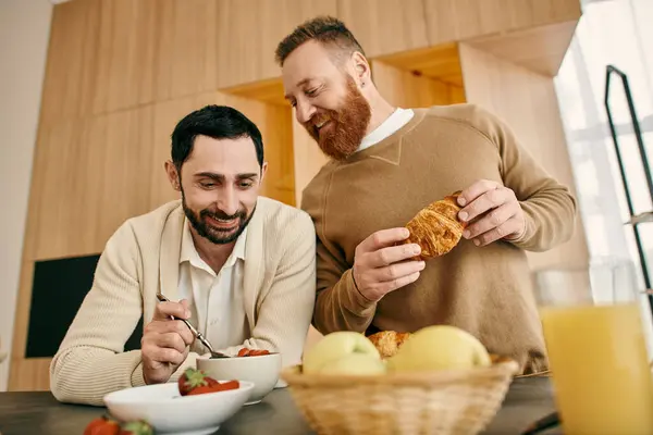 Dois homens felizes, um casal gay, sentam-se em uma mesa de cozinha aconchegante, saboreando café da manhã e companhia uns dos outros em um apartamento moderno. — Fotografia de Stock