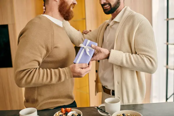 Two men joyfully exchange gift in a modern kitchen, showcasing love and friendship in an intimate setting. — Stock Photo