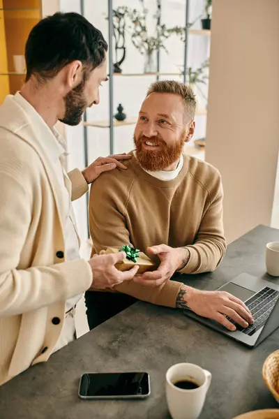 A man happily presents a gift to his partner on a laptop screen in a modern apartment setting. — Stock Photo