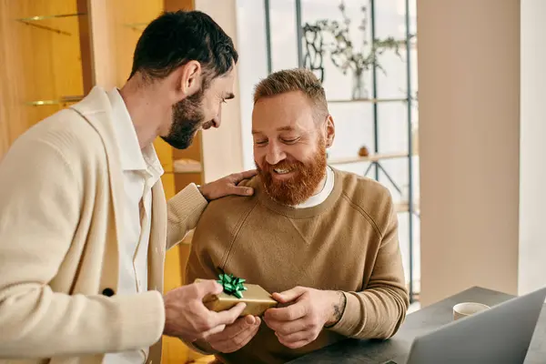 A man joyfully gives a gift to his partner in front of a laptop in a modern setting, symbolizing their shared love and connection. — Stock Photo